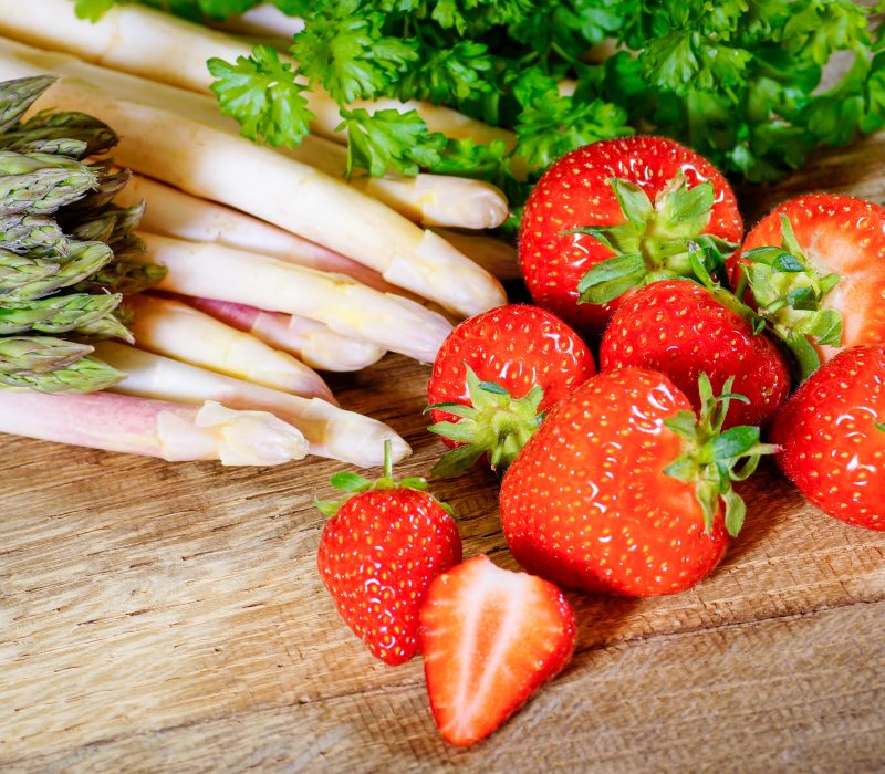 Frischer grüner und weißer Spargel, leuchtend rote Erdbeeren und krause Petersilie auf einer hölzernen Unterlage zelebrieren Spargelsaison und Erdbeersaison mit einem farbenfrohen Arrangement aus Obst und Gemüse.