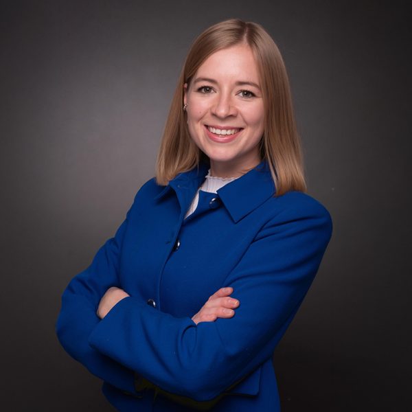 Clara Albrecht, eine Frau mit schulterlangem blondem Haar, steht lächelnd mit verschränkten Armen da. Sie trägt einen blauen Blazer und ein weißes Hemd vor einem dunklen, schlichten Hintergrund.