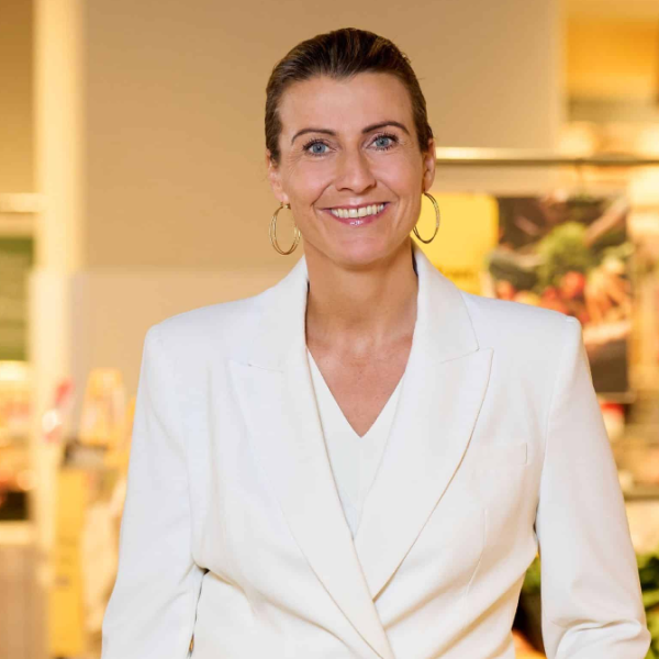 Eine lächelnde Frau in einem weißen Blazer steht drinnen. Sie hat ihr Haar zurückgebunden und trägt große Creolen. Der sanft verschwommene Hintergrund lässt auf eine Einzelhandelsumgebung schließen und verkörpert die Essenz der Regionalität. Könnte sie der bekannte Regionalstar Elisabeth Promberger sein?.
