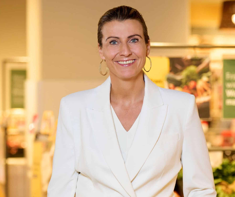 Eine lächelnde Frau in einem weißen Blazer steht drinnen. Sie hat ihr Haar zurückgebunden und trägt große Creolen. Der sanft verschwommene Hintergrund lässt auf eine Einzelhandelsumgebung schließen und verkörpert die Essenz der Regionalität. Könnte sie der bekannte Regionalstar Elisabeth Promberger sein?.