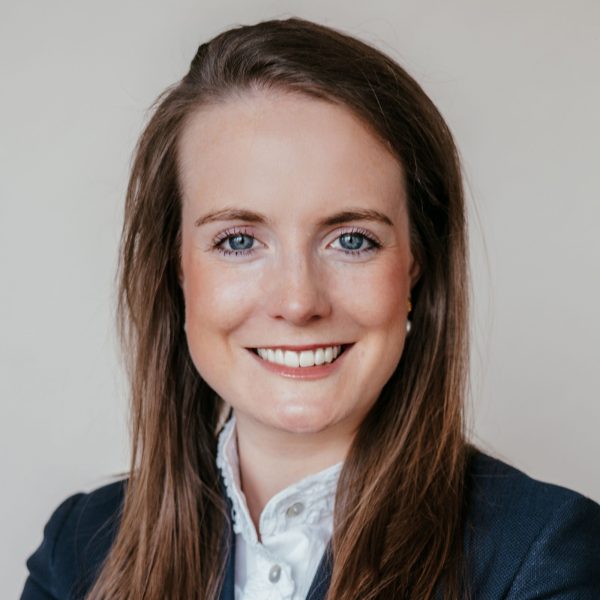 Eine Frau mit langen braunen Haaren, die einen dunklen Blazer und ein weißes Hemd mit Kragen trägt, lächelt in die Kamera vor einem schlichten hellen Hintergrund.