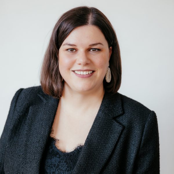 Eine Frau mit glattem braunem Haar und heller Haut lächelt in die Kamera. Sie trägt einen schwarzen Blazer über einem schwarzen Oberteil und baumelnde Ohrringe vor einem schlichten hellen Hintergrund.