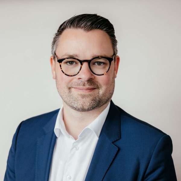 Ein Mann mit kurzen dunklen Haaren, der eine Brille, eine blaue Anzugsjacke und ein weißes Hemd trägt, lächelt leicht in die Kamera vor einem schlichten hellen Hintergrund.