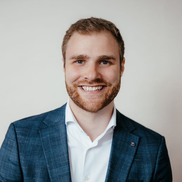 Ein Mann mit hellbraunem Haar und Bart, der eine blaukarierte Anzugsjacke und ein weißes Hemd trägt, lächelt in die Kamera vor einem schlichten hellen Hintergrund.