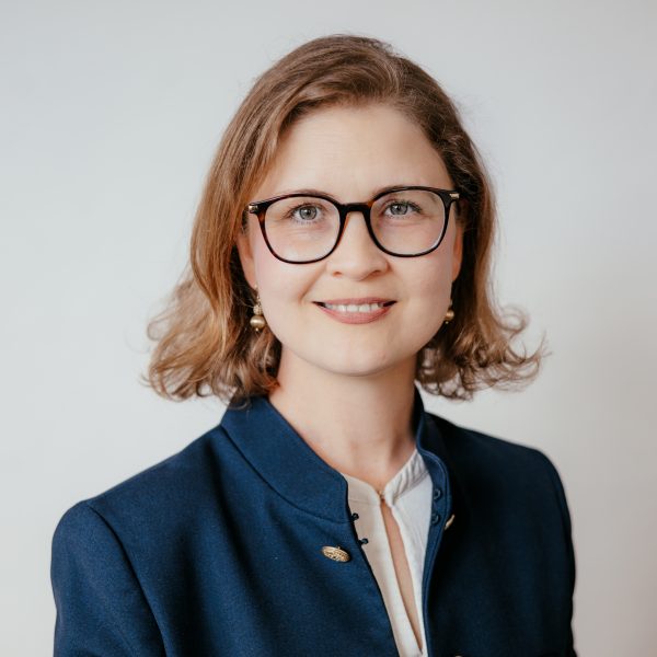 Eine Frau mit schulterlangem hellbraunem Haar, die eine Brille und einen marineblauen Blazer über einer weißen Bluse trägt, lächelt in die Kamera vor einem schlichten hellen Hintergrund.