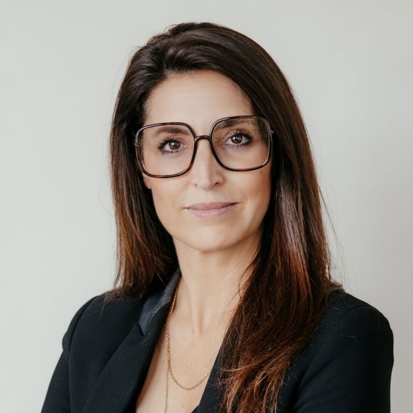 Eine Frau mit langen braunen Haaren, einer großen, eckigen Brille und einem schwarzen Blazer steht vor einem schlichten, hellen Hintergrund und blickt selbstbewusst in die Kamera.