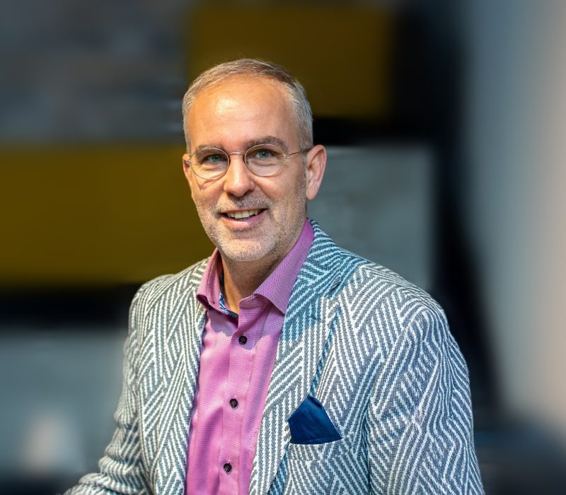 Ein lächelnder Mann mit kurzen grauen Haaren und Brille trägt einen blau-weiß gemusterten Blazer über einem rosa Hemd und steht in einem Haus mit unscharfem Hintergrund.