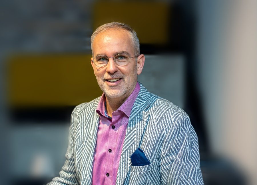 Ein lächelnder Mann mit kurzen grauen Haaren und Brille trägt einen blau-weiß gemusterten Blazer über einem rosa Hemd und steht in einem Haus mit unscharfem Hintergrund.