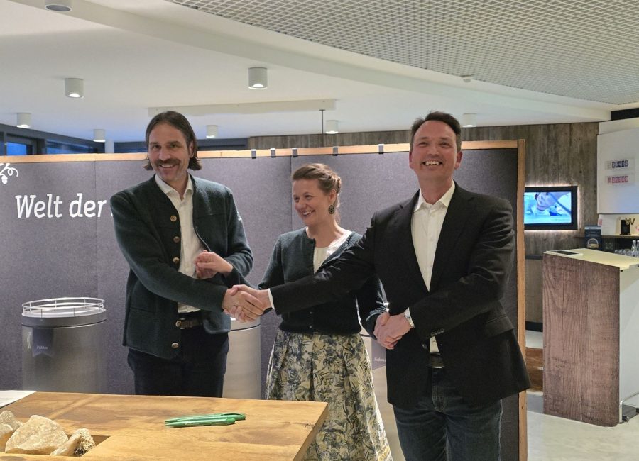 Drei Personen stehen in einem Raum, lächeln und schütteln sich die Hände in einer Dreiecksformation. In der Mitte steht eine Frau, flankiert von zwei Männern. Hinter ihnen steht ein Schild mit der Aufschrift "Welt der- wie ein automatisch gespeicherter Entwurf von Teamarbeit".