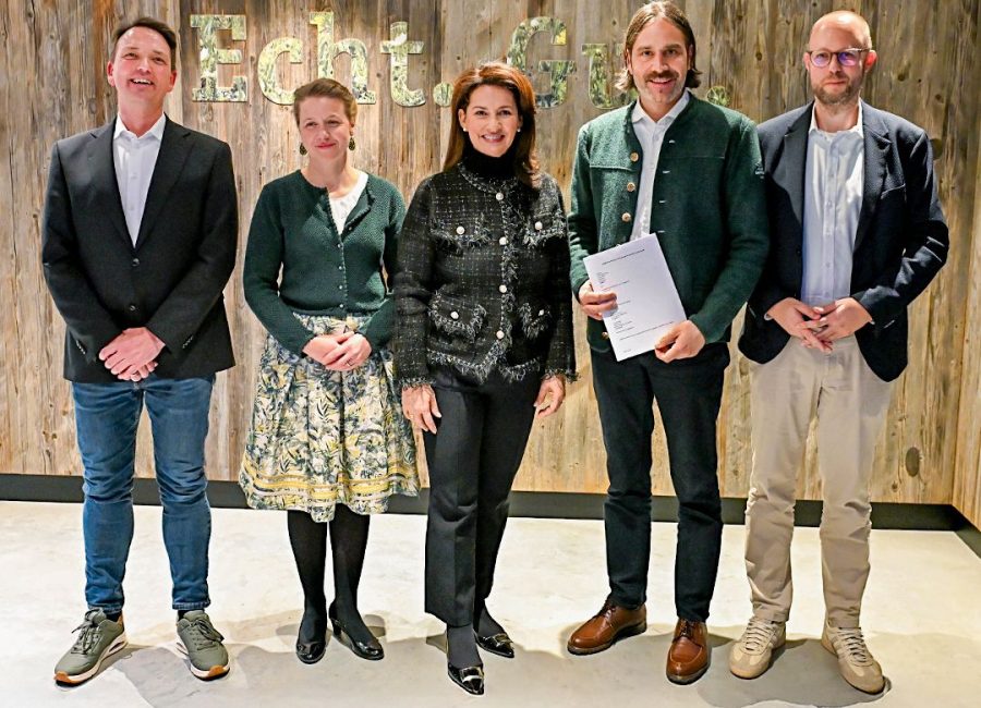 Fünf Personen stehen nebeneinander vor einer Holzwand mit dem Schriftzug "Echt"; drei Männer und zwei Frauen, alle in eleganter oder Business-Casual-Kleidung - ein Mann hält einen automatisch gespeicherten Entwurf.