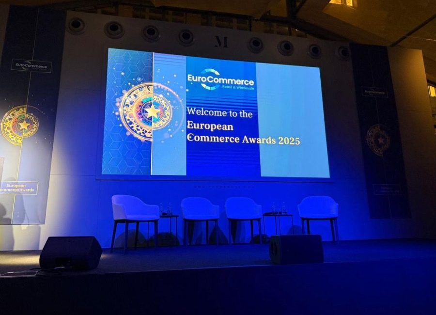 Eine Bühne mit vier weißen Stühlen und zwei kleinen Tischen steht vor einem großen Bildschirm mit der Aufschrift "Welcome to the Commerce Awards 2025" auf der EuroCommerce-Veranstaltung, und blaues Licht erhellt die Szene.