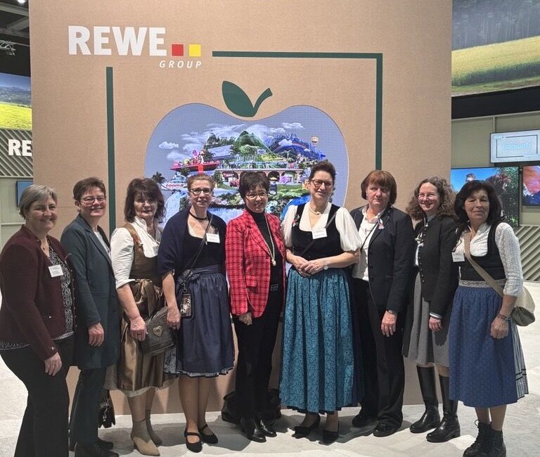 Neun Frauen stehen lächelnd vor einer Auslage der REWE Group mit einer großen Apfelgrafik und überdimensionalen Gemüserequisiten, aufgenommen in einem automatisch gespeicherten Entwurf. Die meisten tragen traditionelle Kleider, eine trägt eine rotkarierte Jacke.