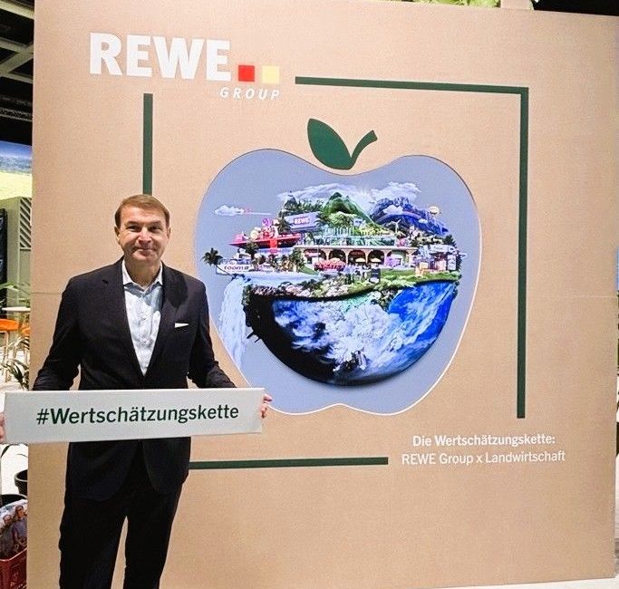 Ein Mann im Anzug steht mit einem Schild mit der Aufschrift #Wertschätzungskette vor einem Display der REWE Group, das einen apfelförmigen Rahmen mit einer Landschaftscollage von Bauernhöfen und Märkten zeigt - ein automatisch gespeicherter Entwurf von Wertschätzung für die Gemeinschaft.