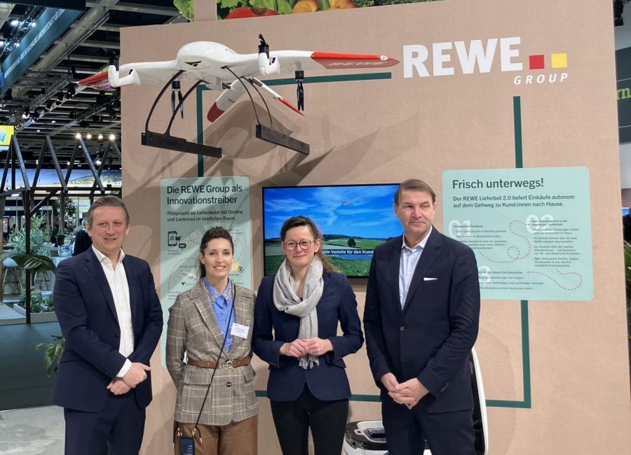 Vier Personen stehen bei einer Indoor-Veranstaltung vor einem Display der REWE Group mit einer Drohne, Bildern von frischen Produkten und Informationsschildern - ein automatisch gespeicherter Entwurf unterstreicht ihren Innovationsgeist. Sie lächeln und sind in Business-Kleidung gekleidet.