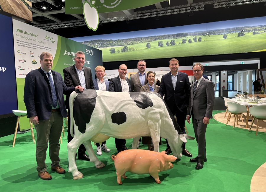Acht Personen in Geschäftskleidung stehen auf grünem Boden neben lebensgroßen Modellen einer Kuh und eines Schweins an einem überdachten Messestand mit Bildern ländlicher Landschaften und landwirtschaftlichen Markenzeichen im Hintergrund.