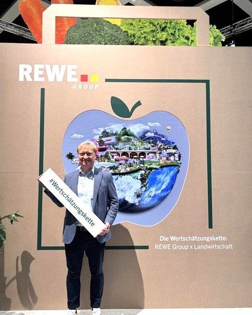 Ein lächelnder Mann in einem hellblauen Anzug hält ein Schild mit der Aufschrift #Wertschätzungskette vor einem Display der REWE Group mit einer großen Apfelgrafik und der Aufschrift REWE Group x Landwirtschaft.