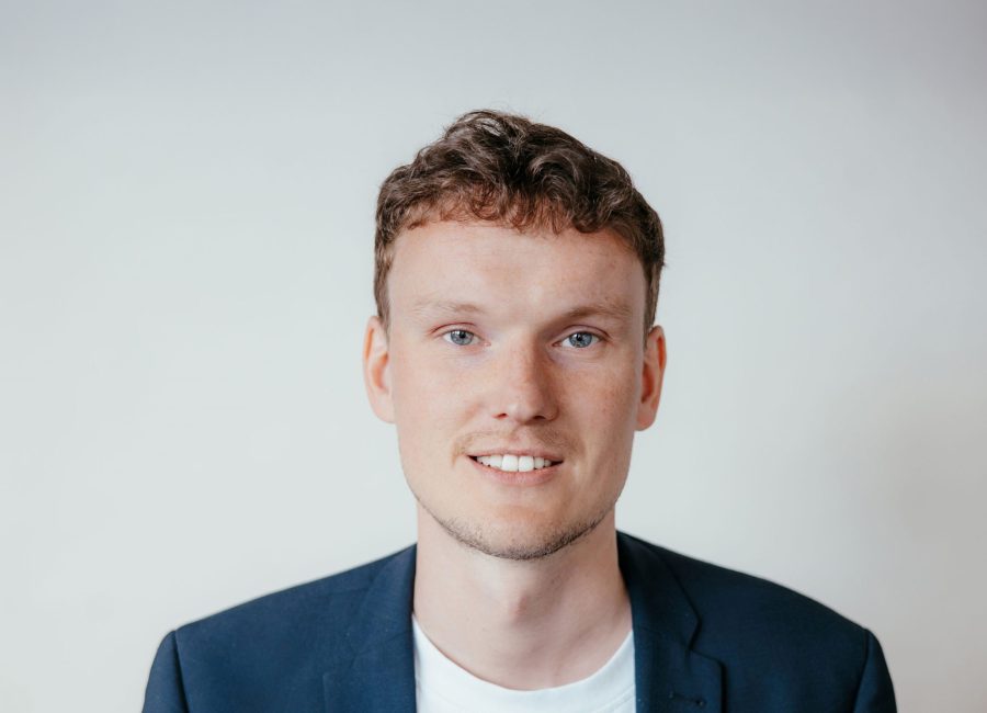 Ein junger Mann mit kurzen, lockigen braunen Haaren und heller Haut lächelt in die Kamera. Er trägt einen dunklen Blazer über einem weißen Hemd und steht vor einem schlichten, hellen Hintergrund.