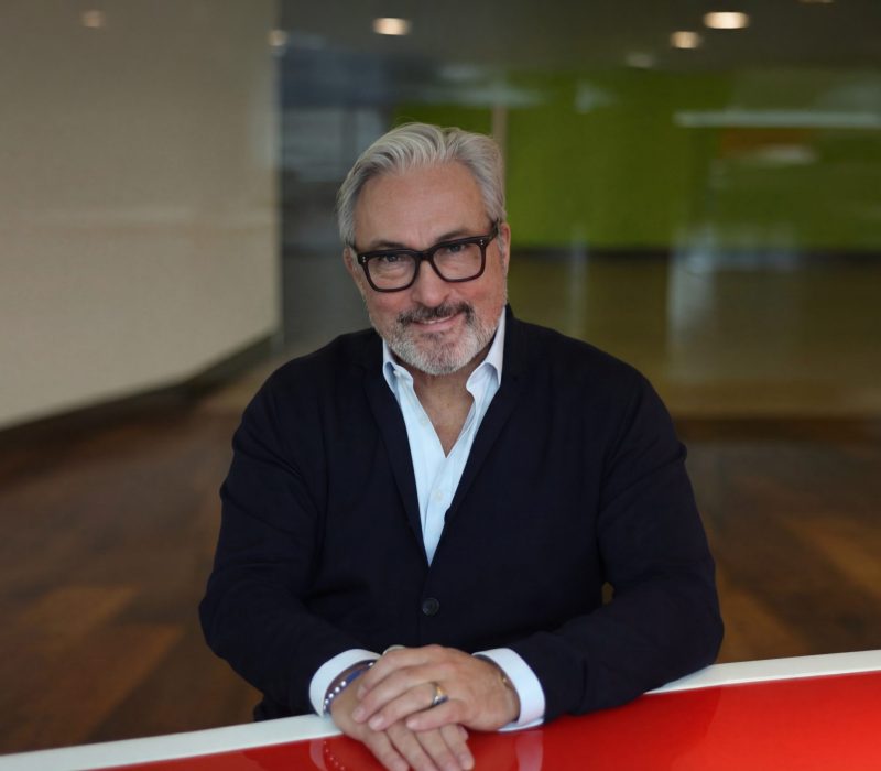 Ein Mann mit grauem Haar, schwarzer Brille und Bart sitzt an einem rot-weißen Tisch, trägt einen dunklen Anzug und ein weißes Hemd und lächelt in einem modernen, hellen Büroraum in die Kamera.