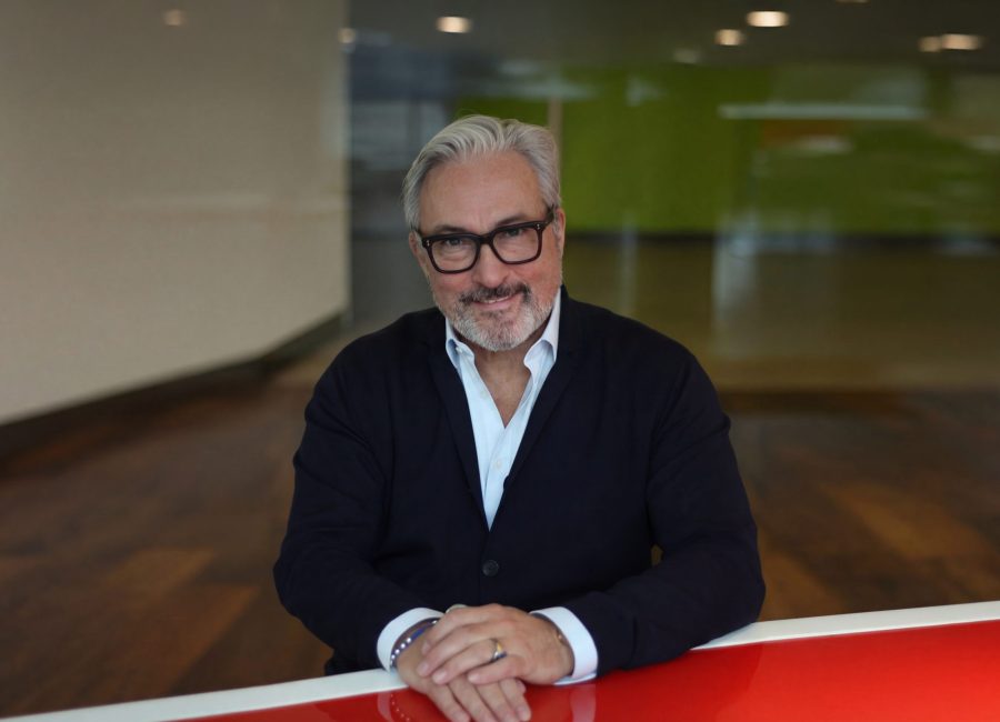 Ein Mann mit grauem Haar, schwarzer Brille und Bart sitzt an einem rot-weißen Tisch, trägt einen dunklen Anzug und ein weißes Hemd und lächelt in einem modernen, hellen Büroraum in die Kamera.