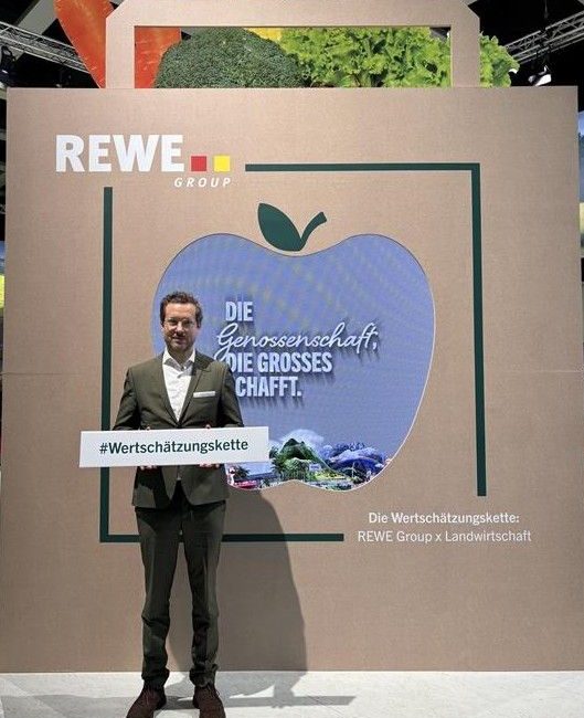 Philipp Hennerkes im Anzug steht vor einem großen apfelförmigen Display mit dem Logo der REWE Group und hält ein Schild mit der Aufschrift #Wertschätzungskette.