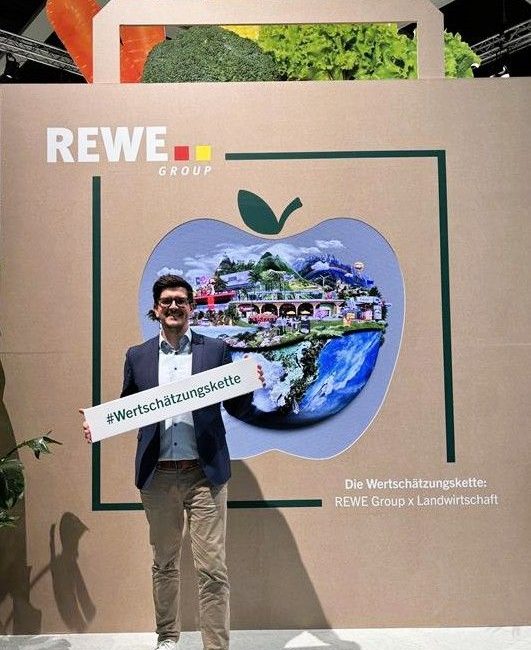 Ein lächelnder Mann mit Brille hält ein Schild mit der Aufschrift #Wertschätzungskette vor einem Display der REWE Group in Form einer Einkaufstasche und eines Apfels mit Lebensmittelbildern und einem deutschen Text, der für Wertschätzung in der Landwirtschaft wirbt - ein automatisch gespeicherter Entwurf.
