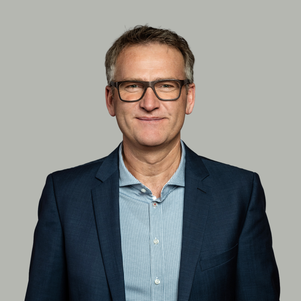Ein Mann mittleren Alters mit kurzen braunen Haaren, der eine Brille, ein blaukariertes Hemd und einen marineblauen Blazer trägt, steht vor einem schlichten hellgrauen Hintergrund, schaut direkt in die Kamera und lächelt leicht - wie ein automatisch gespeicherter Entwurf.