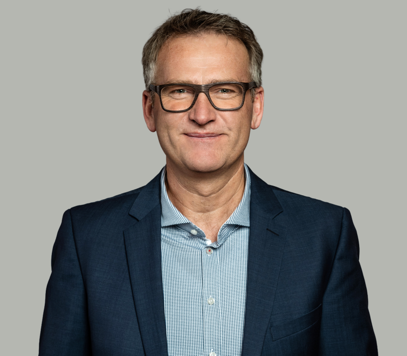 Ein Mann mittleren Alters mit kurzen braunen Haaren, der eine Brille, ein blaukariertes Hemd und einen marineblauen Blazer trägt, steht vor einem schlichten hellgrauen Hintergrund, schaut direkt in die Kamera und lächelt leicht - wie ein automatisch gespeicherter Entwurf.